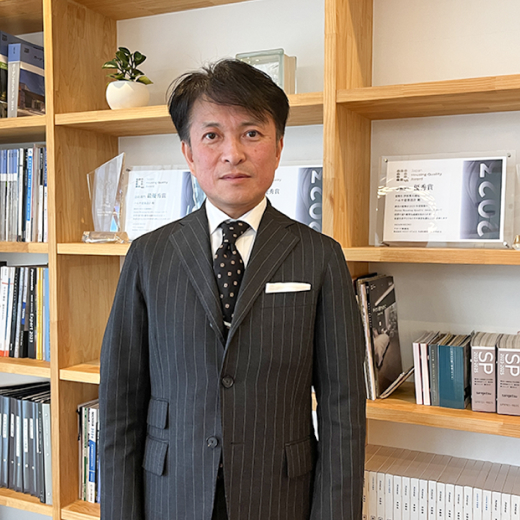 表彰状とトロフィーを置いた棚の前に立つハルサ建築設計のスーツを着た男性
